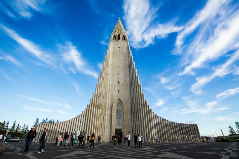 Tourists visiting Hallgr&iacute;mskirkja in Reykjavik, Iceland, a prominent landmark with a striking modern architectural design under a clear blue sky.