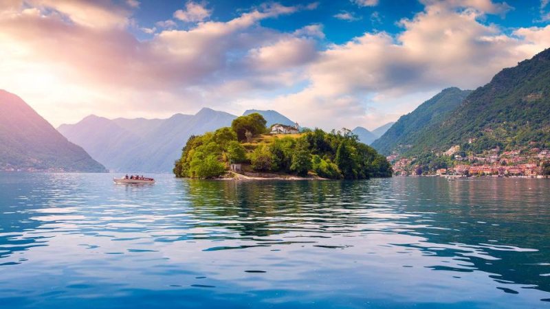 Scenic view of Lake Como in November with tranquil water and an island surrounded by autumn foliage. A small boat glides across the lake under a partly cloudy sky, with mountains in the background. Perfect for travel tips focused on less crowded visits and enjoying fall landscapes.