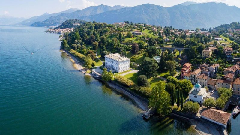 Aerial view of a picturesque villa and lush greenery by the shores of Lake Como with mountains in the background, showcasing a serene autumn landscape in November.
