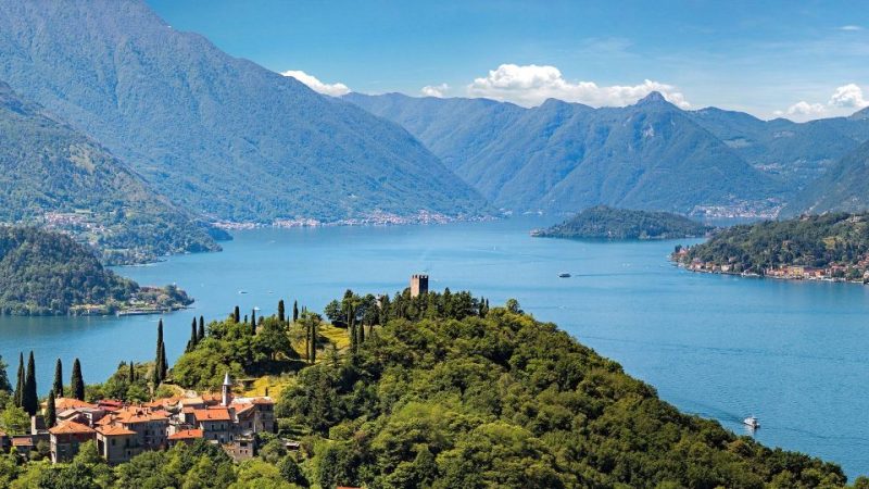 Panoramic view of Lake Como with lush green hills, historic buildings, and autumn foliage under a clear blue sky, showcasing the serene beauty of this popular travel destination in November.