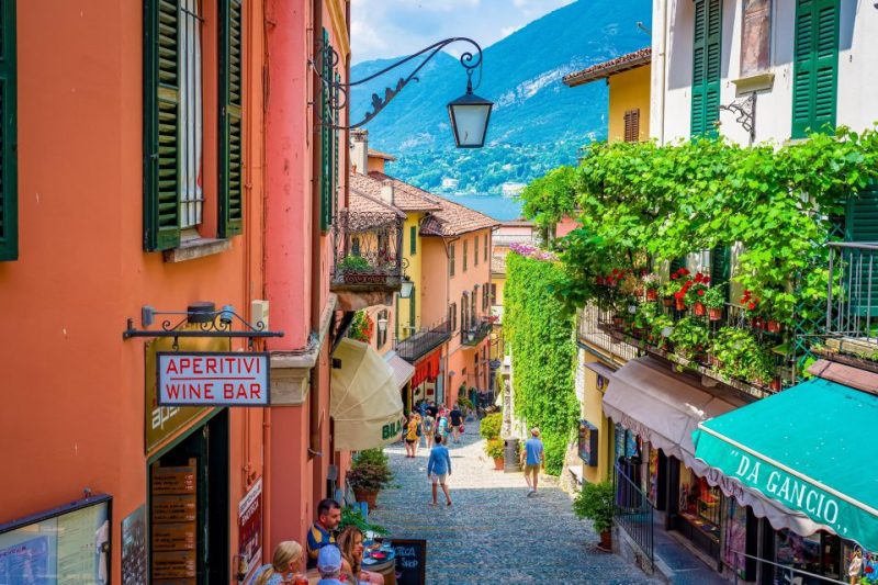 Colorful street in Lake Como with vibrant orange and yellow buildings, a wine bar sign, and lush green plants. Tourists stroll along the cobbled path enjoying the scenic view with mountains in the background, capturing the essence of a peaceful November visit.