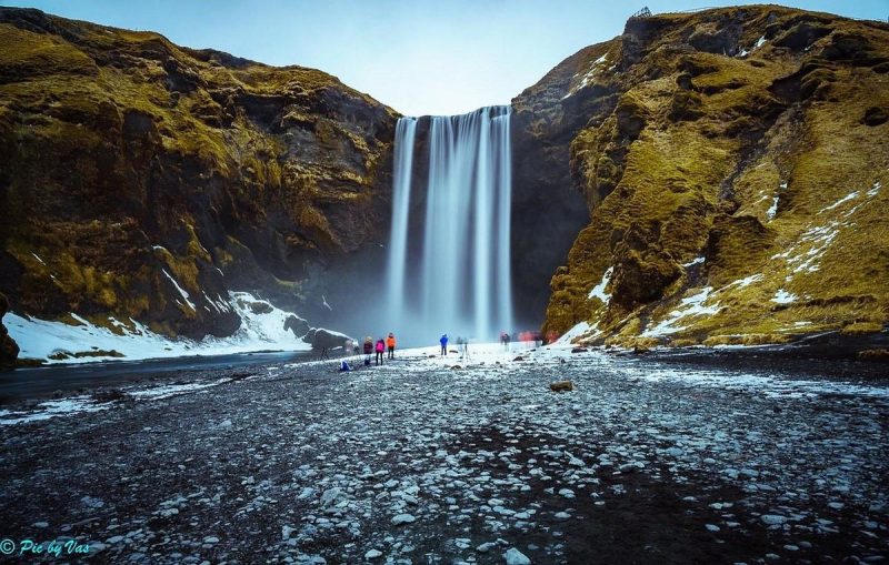 Majestic Sk&oacute;gafoss waterfall in Iceland, surrounded by rugged cliffs and partially frozen ground, with tourists admiring the view.