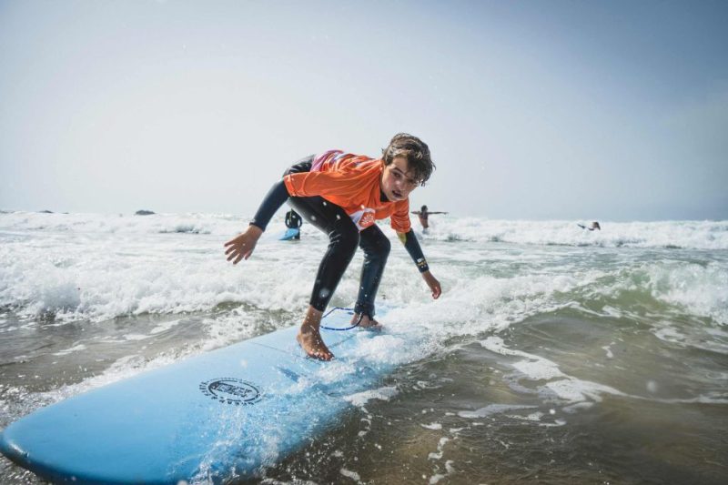 Young surfer in orange shirt riding a wave on a blue surfboard in Kona, Hawaii, showcasing popular water activities and attractions.