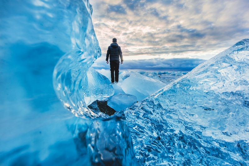 Person standing on glacial ice at Jokulsarlon Glacier Lagoon in Iceland, surrounded by large blue icebergs with a dramatic cloudy sky, highlighting the natural beauty and adventure of exploring Iceland's landscapes.