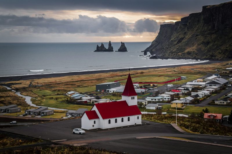 Scenic view of Vik village in Iceland, featuring a church with a red roof in the foreground, surrounded by colorful houses and dramatic cliffs along a black sand beach, with the Reynisdrangar sea stacks in the distance under a cloudy sky.