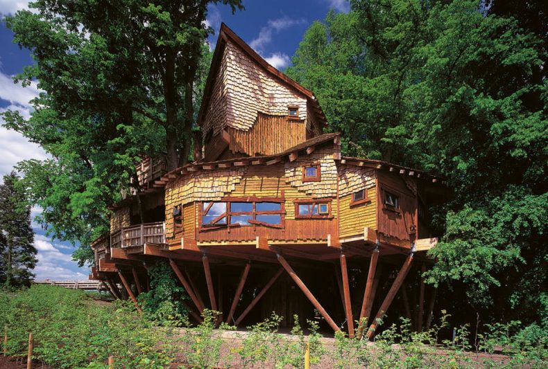 Unique wooden treehouse in Alnwick Garden, UK, featuring rustic architecture with shingled roof and supported by stilts, surrounded by lush greenery.