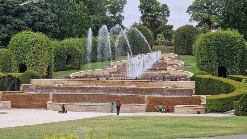 View of Alnwick Poison Garden's grand water cascade feature with people walking and cycling, surrounded by lush, sculpted hedges and trees.