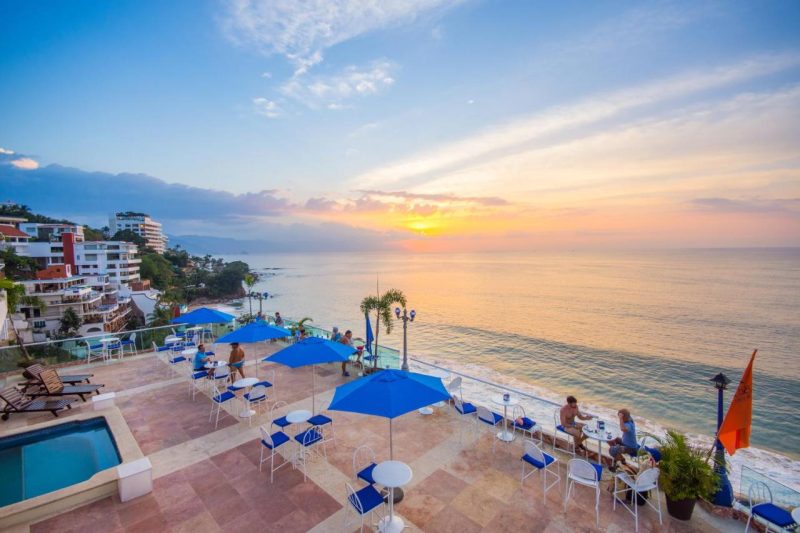 Scenic view from a terrace at a clothing optional resort in Puerto Vallarta, featuring a stunning sunset over the ocean, with blue umbrellas and outdoor seating for relaxation.