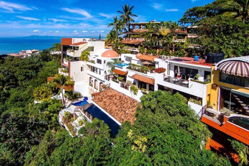 Aerial view of a luxurious resort surrounded by lush greenery and overlooking the ocean in Puerto Vallarta, featuring vibrant architecture and open terraces.