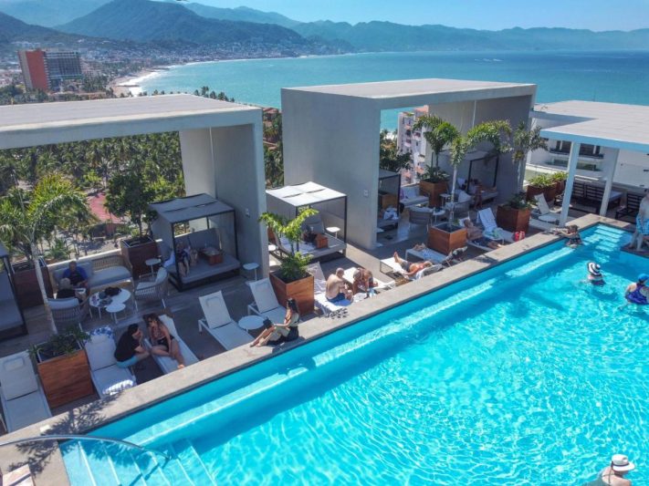 Aerial view of a luxurious rooftop pool at a resort in Puerto Vallarta, featuring lounge chairs, cabanas, and palm trees with a stunning ocean view in the background.