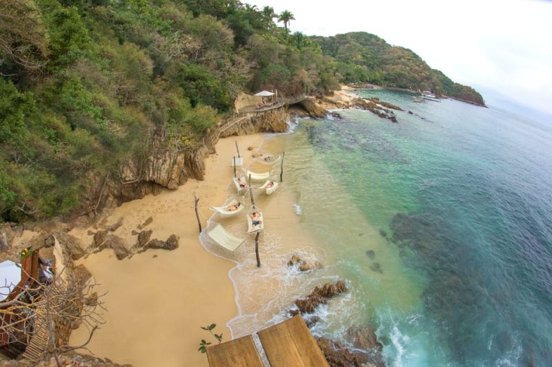 Aerial view of a secluded beach with clear turquoise water, surrounded by lush green vegetation and rocky cliffs in Puerto Vallarta. The sandy shore features four hammocks suspended over the sand, perfect for relaxation at a clothing-optional resort.