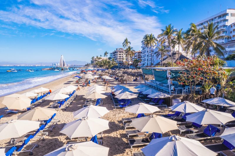 Sunny beach scene in Puerto Vallarta featuring numerous beach umbrellas and lounge chairs on sandy shore, surrounded by palm trees and nearby resort buildings, with a vibrant blue sky overhead.