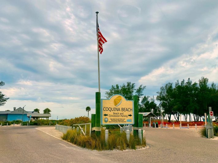 Coquina Beach entrance with parking sign and American flag under cloudy sky in Sarasota, Florida.