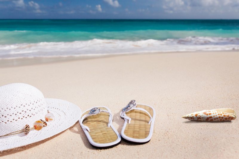 White sun hat, bejeweled flip-flops, and a conch shell on a sandy beach with turquoise water in Sarasota, Florida.