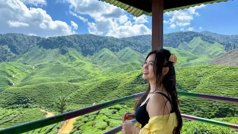 Woman enjoying scenic view of lush green tea plantations in Cameron Highlands, Malaysia, under a blue sky; perfect for a short vacation or getaway.